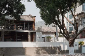 A serene Asian-style building with a white exterior, surrounded by trees, featuring a balcony and large window, with chairs placed outside