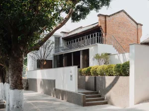 A modern house with a white facade and a red brick wall, featuring a patio, balcony, and staircase, surrounded by trees