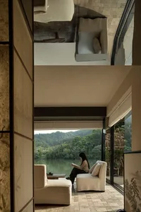A serene living space with a modern design, featuring a woman sitting on a white chair in front of a window overlooking a lake, with a couch and another chair nearby
