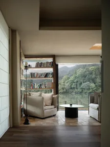A bright and inviting living space with a large window overlooking a lake, featuring two couches, a chair, and a coffee table, surrounded by books and filled with natural light