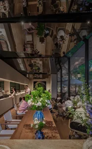A modern dining space with wooden floors, artwork on the walls, and natural light from windows, featuring a large table and smaller tables with people seated and engaged in conversation