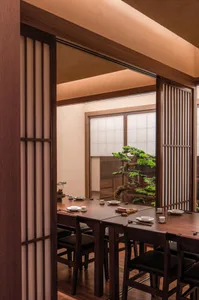 Cozy traditional Japanese dining space with a wooden table, chairs, and a potted plant, featuring sliding doors and a tatami mat area, well-lit through natural light