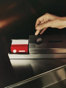 A person's hand places a coin into a metal box with a red tint in a public indoor environment