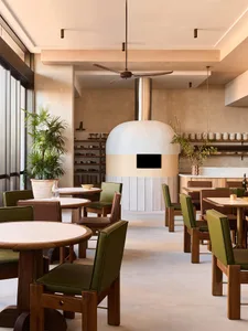 Cozy modern restaurant dining area with a stone oven, wooden tables, and vases, surrounded by chairs and warm lighting