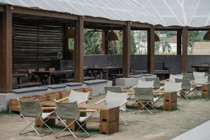 A restaurant patio area with wooden tables and chairs under a large umbrella, set on a dirt ground with a garden-like atmosphere
