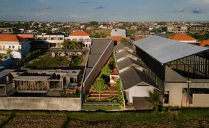A modern hotel and house connected by a bridge, surrounded by potted plants and chairs, with a concrete exterior and inviting atmosphere