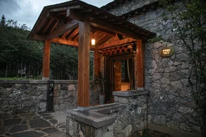 A traditional Asian-style house with a stone exterior, wooden roof overhang, and potted plants, set in a rustic living environment