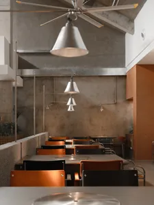 Empty restaurant interior with multiple dining tables and chairs, concrete and wood walls, and overhead lighting