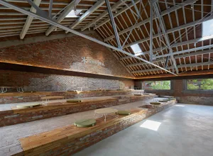 A large brick building with a high ceiling, featuring a dining area with multiple tables and chairs, and several benches for social gatherings or events