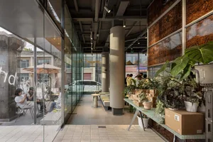 Bright and inviting cafe with glass walls, wooden furniture, and potted plants, featuring multiple dining tables and people socializing