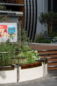 Outdoor seating area with wooden and metal benches, dining tables, and potted plants in a restaurant or cafe setting