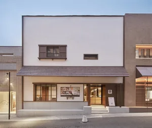 A modern building with a white exterior, brown roof, and wooden door, featuring a clock on the wall and two chairs in the scene