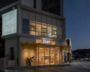 A modern restaurant with a glass storefront, lit interior, and potted plants, set at night with evening atmosphere