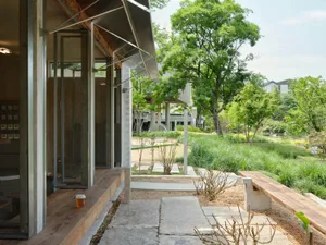 A serene outdoor space with a modern building featuring concrete walls, a metal roof, and a patio area with a bench, dining table, and potted plants