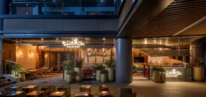 A modern Asian restaurant with a glass building, black roof, and potted plants, featuring a dining area with chairs and a TV