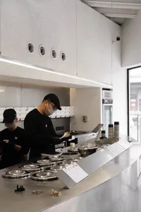Industrial kitchen with two people working, wearing face masks, surrounded by appliances, bowls, and cups