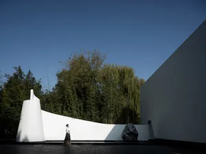 A woman stands in front of a modern building with a mirror, surrounded by a calm and serene environment with two large rocks nearby