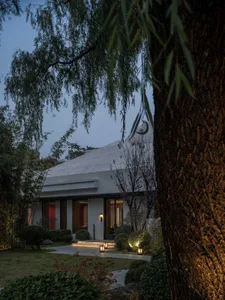 A modern building with a white exterior, a balcony, and a tree in front, surrounded by potted plants and a bench
