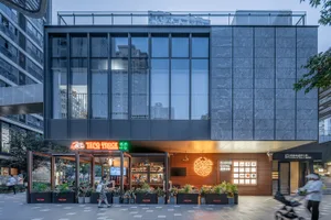 Modern restaurant with a glass front, wood accents, and metal elements, featuring potted plants, tables, chairs, and a TV, creating a stylish and inviting atmosphere