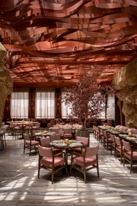 A warm and inviting restaurant interior with multiple tables, chairs, potted plants, and a unique tree-inspired wall design
