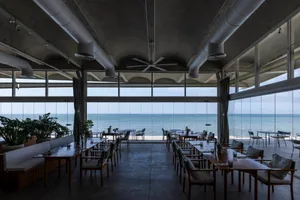 Dining area with multiple tables and chairs, large window overlooking the ocean, and a relaxed atmosphere