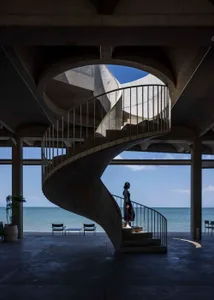 A modern spiral staircase leads to a balcony overlooking the ocean, with two chairs on the ground near the bottom of the stairs