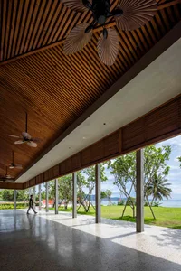 Warm and inviting open-plan space with wooden beams, a large window, and natural light, featuring a living area and dining space with chairs and a person walking through