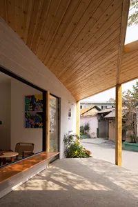 Modern house with wooden walls, a white roof, and a patio area with a bench and dining table, surrounded by chairs and a living room with a couch and potted plant