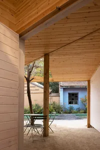 A calm and inviting outdoor patio with wooden walls, a green table, and two chairs, set in front of a blue building