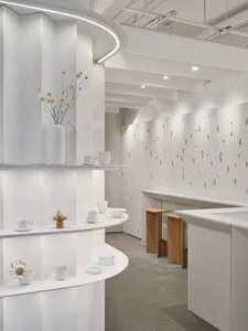 A well-lit kitchen showroom with white surfaces, a sink, countertop, and decorative items such as cups, bowls, vases, and potted plants