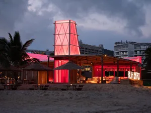 An open-air restaurant on a beach at night with a modern wooden building and red roof, surrounded by chairs and a dining table