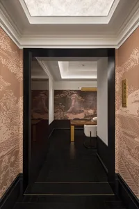 Asian-inspired living room with a wooden table, chairs, and couch, adorned with intricate artwork and a black doorway leading to another room