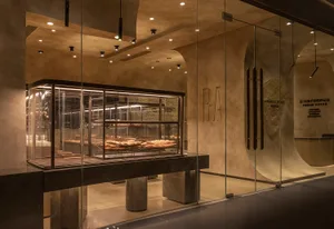 A modern restaurant interior with a glass display case showcasing sandwiches and cakes, surrounded by neutral-toned walls and a minimalist aesthetic