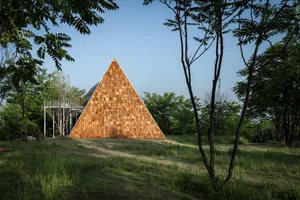 A unique wooden house with a triangular shape, surrounded by trees and grass in a serene natural setting