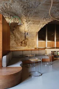 A modern living and dining area with two couches, three chairs, two dining tables, and a stone wall, lit by natural light, with books and a welcoming atmosphere