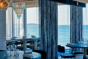 Elegant dining space with modern design, blue walls, and a large window offering ocean views, featuring tables, chairs, and a bar with wine glasses