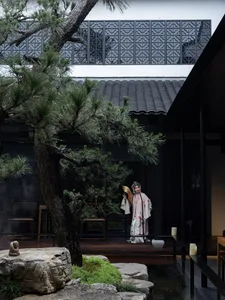 A serene courtyard setting with a woman in traditional attire standing in front of a building, surrounded by trees and several chairs, conveying a sense of cultural significance and tranquility