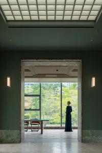A woman stands by a window overlooking a garden, surrounded by a dining table and chairs in a comfortable and inviting living space