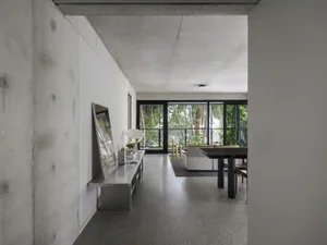 Open modern living space with concrete walls, wooden floors, and natural light from a large window, featuring a kitchenette, dining tables, and potted plants