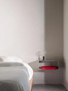 A simple white bedroom with a bed against a wall, a small shelf with two books, one red, in a clean and bright space