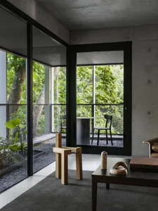 Modern living space with concrete walls and flooring, featuring a large window overlooking a forest, two chairs, a dining table, and a potted plant
