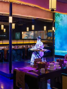 A woman enjoys her meal at a table in front of a large painting in a cozy Asian restaurant with multiple tables and chairs, cups on surfaces, and a relaxed atmosphere