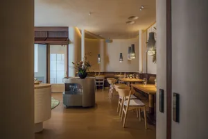 Warm and inviting restaurant interior with wooden floors, tables, chairs, and potted plants, featuring a kitchen in the background