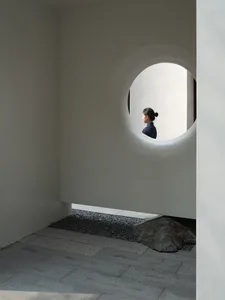 A woman stands in front of a concrete wall with a round window, surrounded by a calm and contemplative atmosphere