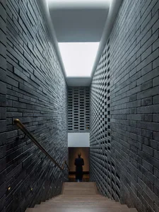 A dimly lit hallway with a woman walking down stone stairs, flanked by a brick wall