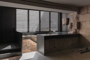 Modern kitchen with a black countertop, stone wall, and natural light from a window, featuring two sinks and a decorative vase