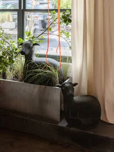 An indoor space with two black sheep statues in a metal pot with grass, near a window with natural light and potted plants