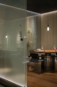 Modern dining space with a glass partition separating the kitchen from the dining area, featuring two chairs and a table set for a meal, with a smaller dining table in the background