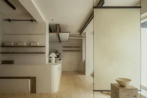 Empty minimalist kitchen dining space with a white wall, brown brick flooring, and a dining table near the center, with bowls and bottles scattered around