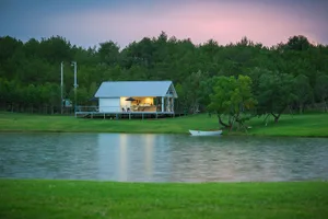 A serene lakeside house with a white exterior, a deck, and a boat docked nearby, surrounded by a grassy area